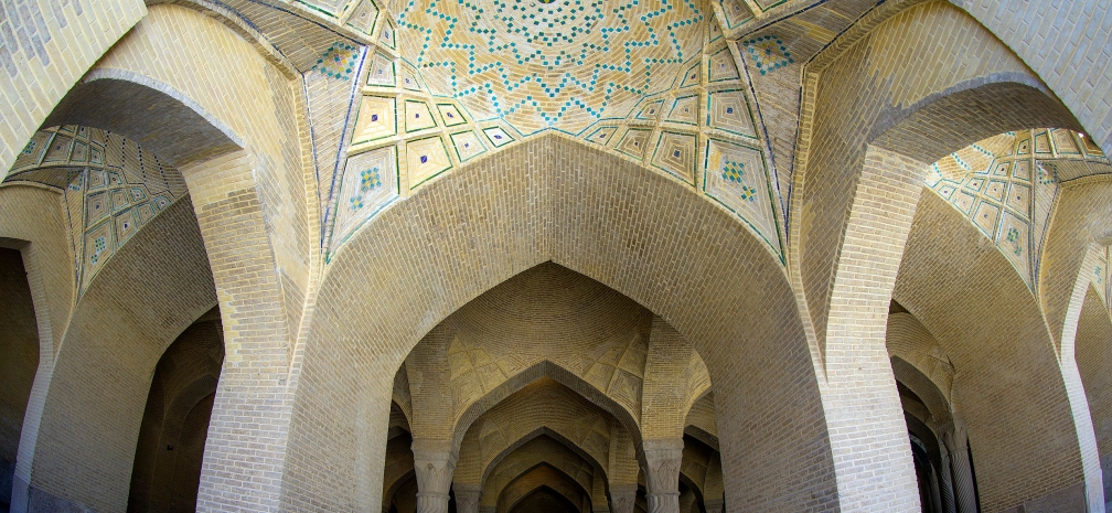 Mostafa Meraji, Vakil Mosque in Shariz, Iran. Big faded brick pillars recede into a dark open hall with an ornately blue and yellow-tiled arching ceiling.