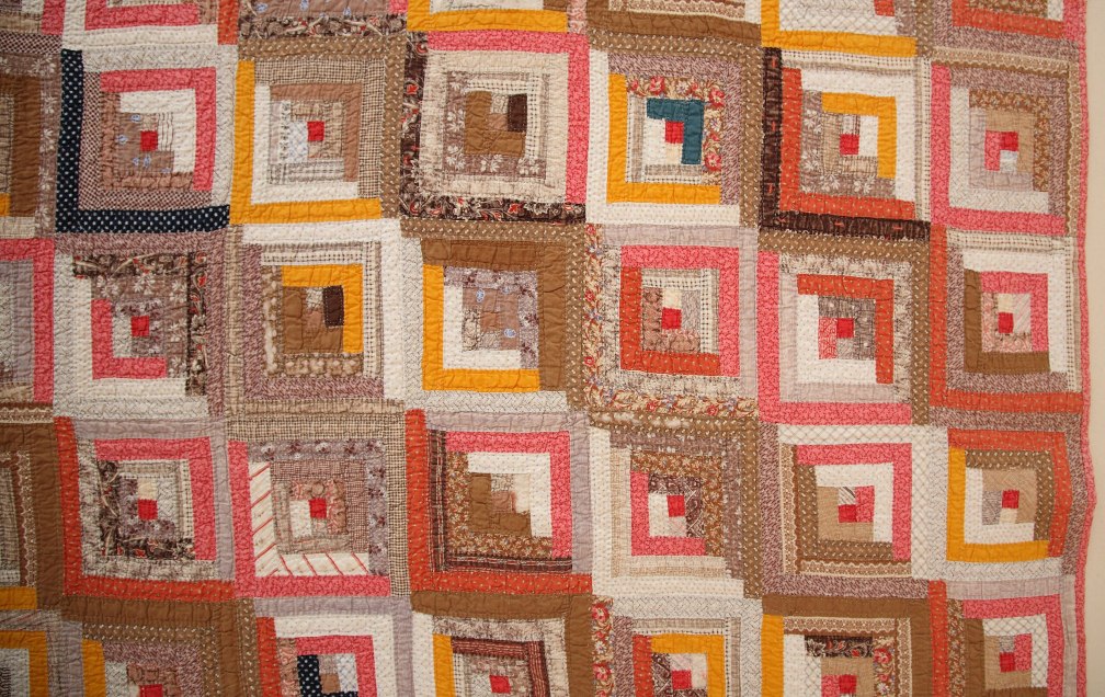 The photo is a close up of a quilt of made up of concentric squares of browns, reds, and yellows. The fabric coloring gives the impression of diagonal bands of dark and light from top left to bottom right.