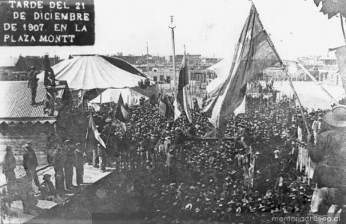 An image of a few thousand people standing in an open square with large flags being held up and waving in the wind. People are standing on the roofs of buildings looking out over the crowd.