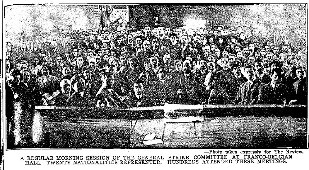 Image of a crowd of people in a large meeting hall all facing the camera, presumably posing for a picture. A caption underneath reads "A regular morning session of the general strike committee at Franco-Belgian Hall. Twenty nationalities represented. Hundreds attended these meetings."