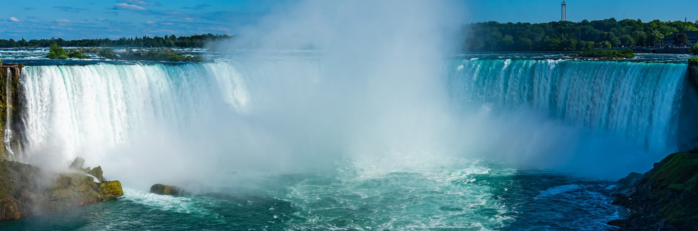 A wide shot of the Niagara Falls horseshoe waterfall. The sun is shining and the water is as blue as the sky.