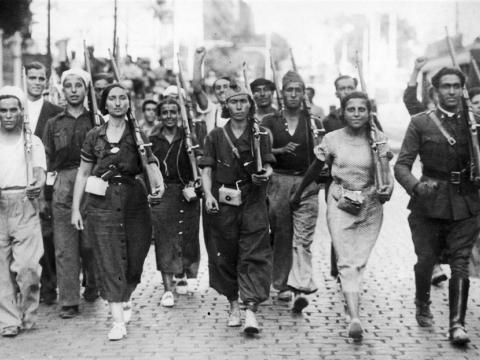 A couple dozen men and women with rifles in their arms are shown marching down the street and toward the camera. Some look happy and others are sober.